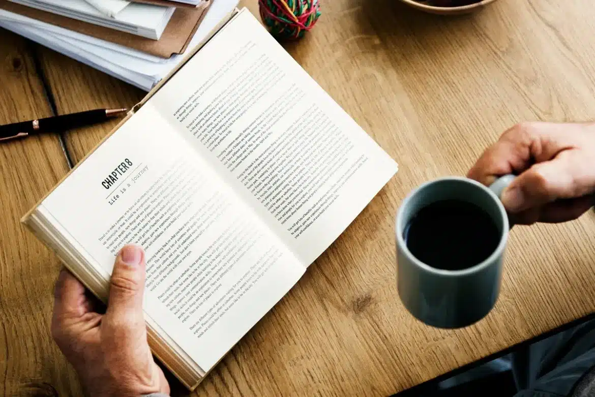 Un livre ouvert avec une tasse de café et des lunettes de lecture sur une table en bois, baignés de lumière naturelle.